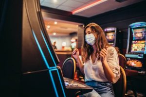 Beautiful young woman with protective face mask playing slot machine in casino.